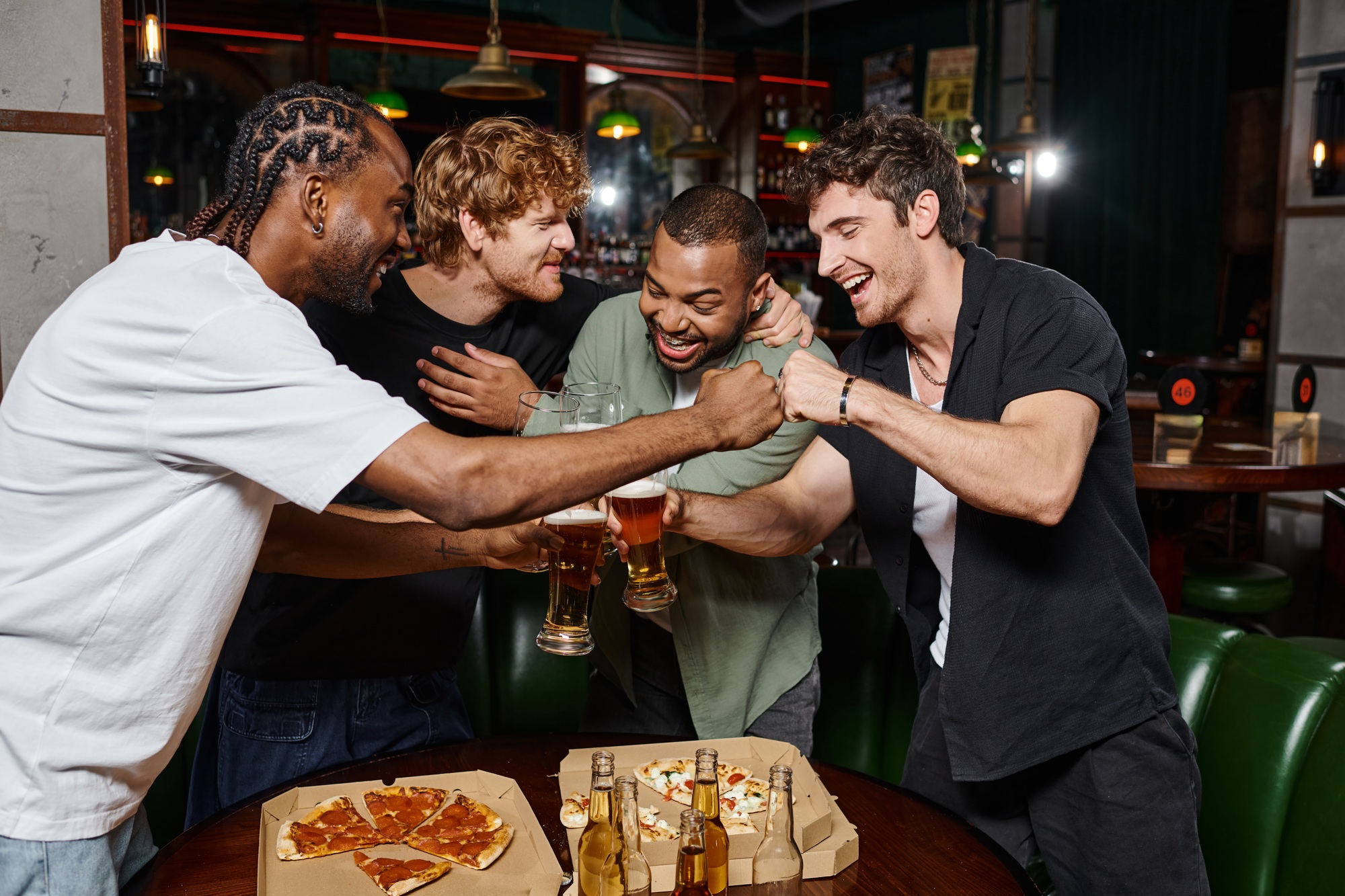 happy interracial male friends fist bumping while toasting glasses of beer in bar, bachelor party